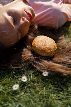 Successful business woman lying on grass with fast food burger cheesburger on hair enjoying leisure...