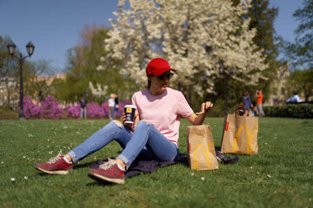 RIGA, LATVIA - APRIL 28, 2019: Successful business woman eating McDonalds Big Mac burger cheesburger and drinking Coca Cola enjoys her leisure free time in a parkのeditorial素材