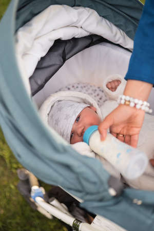 Mother feeds baby boy toddler child milk from a plastic bottle with a bright blue nipple - Caucasian white child with a parents hand visible - Laying in a baby carriage with white sheetsの写真素材