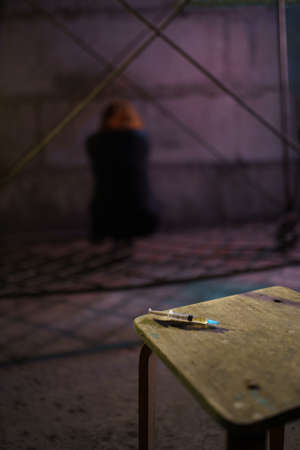 Syringe close up on table with a drug addiction redhead caucasian white young woman in the background sitting and walking away wearing black sweater, skirt and chockerの写真素材