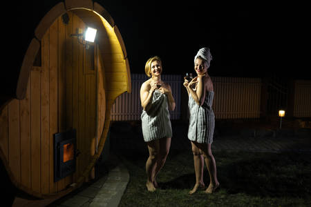 Two girlfriends women drinking red wine from glasses outsite near a wooden mobile sauna wearing just towelsの写真素材