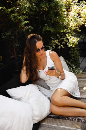 Beautiful woman in white sitting in an outdoor cafe garden busy using texting in mobile phoneの写真素材