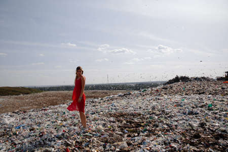 Nature pollution activist at a huge trash dump outdoors - Young blonde woman in a red dress -...
