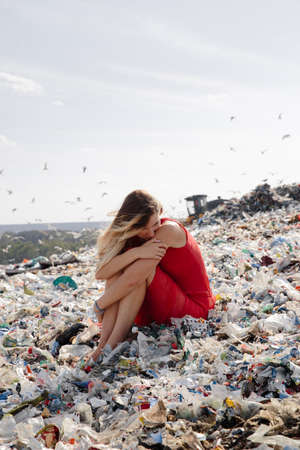 Nature pollution activist at a huge trash dump outdoors - Young blonde woman in a red dress - Looking at all the human waste and plastic in our world - Seagulls flying in Eastern Europe Latvia Rigaの写真素材