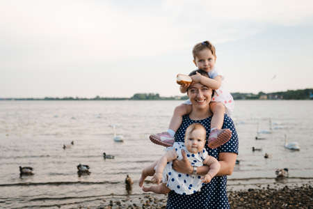 Young mother with her baby girl daughters feeding swan and little ducklings birds bread at a river wearing dotted dress - Family values warm color summer sceneの写真素材