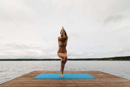 Fit slim woman practicing yoga exercises wearing mini swimsuit bikini at lake with clouds - Yoga meditation and wellness lifestyle concept - European Eastern Latvia Riga.の写真素材