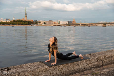 Yoga meditation and wellness lifestyle concept. Fit slim woman practicing yoga exercises near river Daugava and Vecriga old town background at sunset - Full sitting shot and meditating on rock beachの写真素材