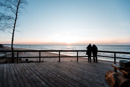 Couple watch sunset at Saulkrasti white dune with sea and sky background.