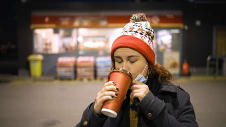 Poor woman lowers medical mask to drink coffee wearing Xmas hatの写真素材