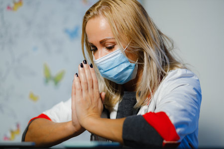 Exhausted female doctor in medical mask praying during home visitの写真素材