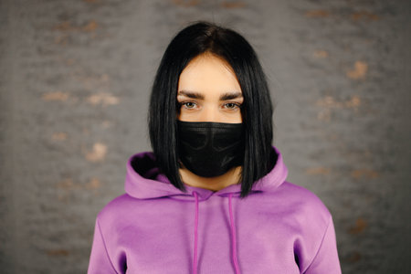 Serious young woman in black protective mask looking at you over home wall backgroundの写真素材