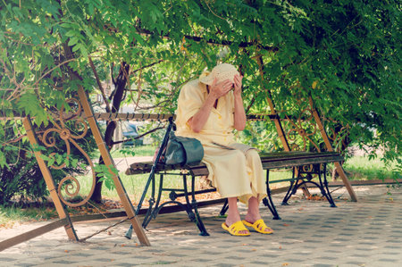 Senior woman sitting on park bench, reading a newspaper. Lonely elderly woman sitting on a bench in summer parkの写真素材