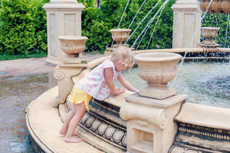 Near The Fountain. Girl having fun with fountain at hot sunny day. Cute little girl playing with fountain splashの写真素材