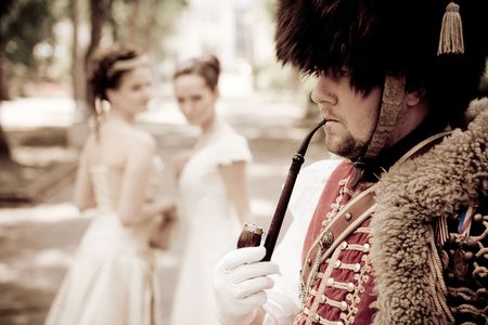 Russian hussar in vintage outfit is smoking his pipe. Two pretty women on the background are looking...