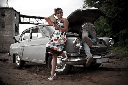 Pretty couple is trying to repair their old car using manual. Rural background.