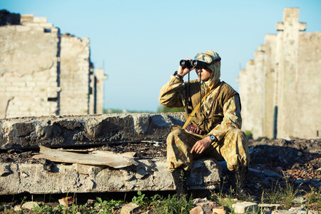 Man in camouflage uniform is looking through the binoculars. Urban ruins on the background.の写真素材