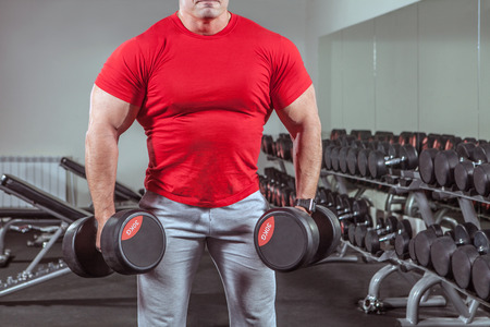 Powerful man with muscular build is training with the dumbbells in the gymの写真素材