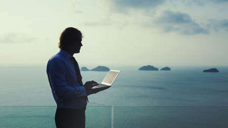 Blond businessman is working on laptop on the tropical ocean background. Artistic toning.の写真素材