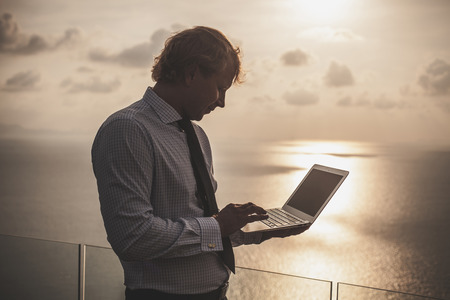 Blond businessman is working on laptop on the tropical ocean background. Artistic toning.の写真素材