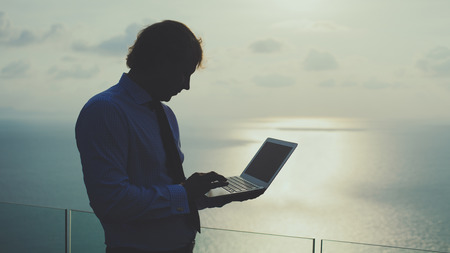 Blond businessman is working on laptop on the tropical ocean background. Artistic toning.の写真素材