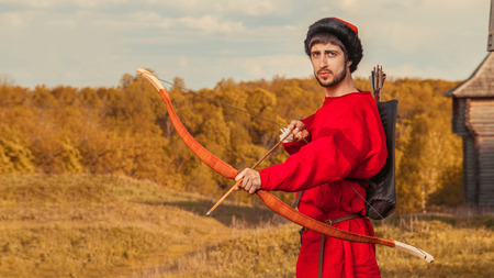 Russian archer with the wooden bow in traditional red suit and fur hat. Summer forest on the...