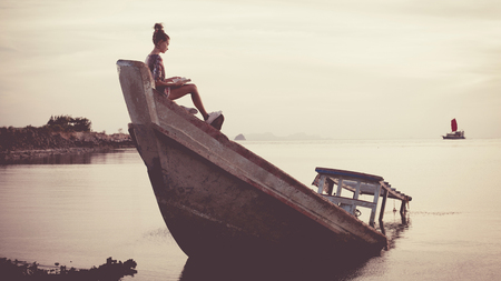Young woman is sitting on the shipwreck and reading a book. Low contrast post-processing.の写真素材