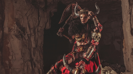 Beautiful woman warrior dressed in steel armor is holding an axe and skull. Dungeon on the background.の写真素材