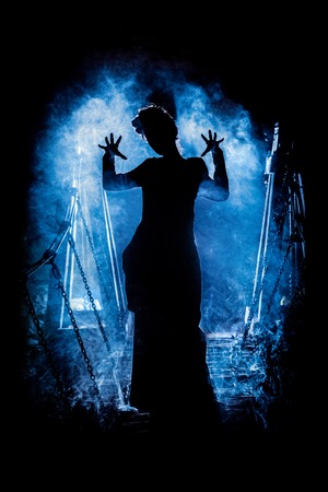 Silhouette of a mysterious witch saying the spell and stretching her hands to us, with clouds of smoke around her, and dungeon bridge on the backgroundの写真素材