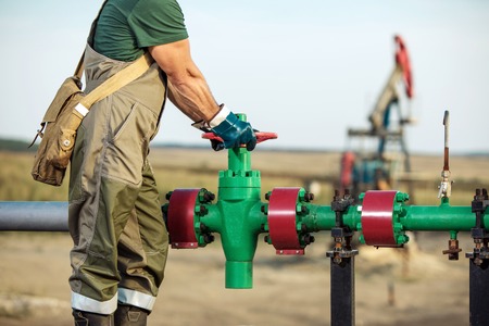 Oil worker is turning valve on the oil pipeline, oil deposit on the background.