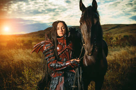 Girl in medieval knight's armor with a horse against the sunset fields backgroundの写真素材