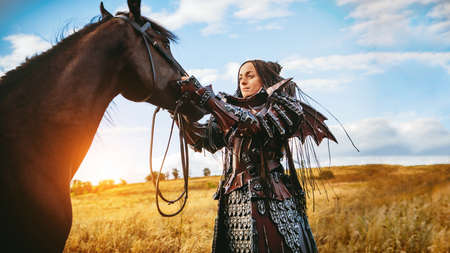 Girl in medieval knight's armor with a horse against the sunset fields background