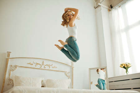 happy young woman jumping on the bed. young blonde woman jumping on the bed and raises his hands to the top. curly blonde jumping on the bed.Cheerful young woman jumping on the bed at homeの写真素材