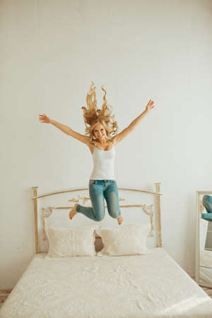 happy young woman jumping on the bed. young blonde woman jumping on the bed and raises his hands to the top. curly blonde jumping on the bed.Cheerful young woman jumping on the bed at homeの写真素材