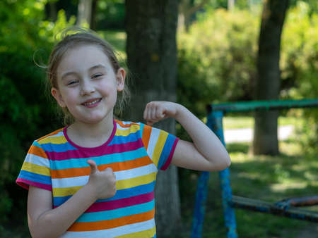 Outdoor sports ground with exercise equipment. Little girl 5-6 years old after training on simulators. She smiles and demonstrates her biceps on her right hand and with a sign that she is doing well.の写真素材
