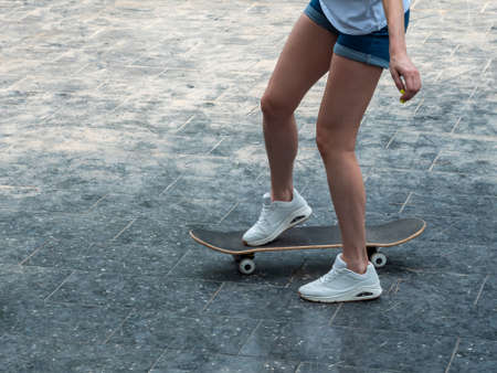 Close up photo of cool young girl with beautiful legs in white sneakers riding skateboard in shiny...