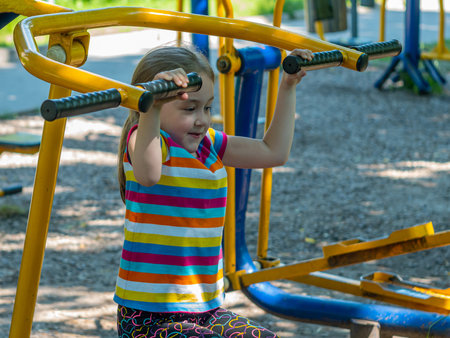 Sports ground in the city park with fitness equipment. Girl 5-6 years old uses a simulator to pump up the muscles of the arms.の写真素材