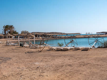 View of empty beach - white deck chairs and abandoned sun umbrellas near seawater, palm trees, and a bungalow on the horizon. The landscape of an abandoned summer resort.の写真素材