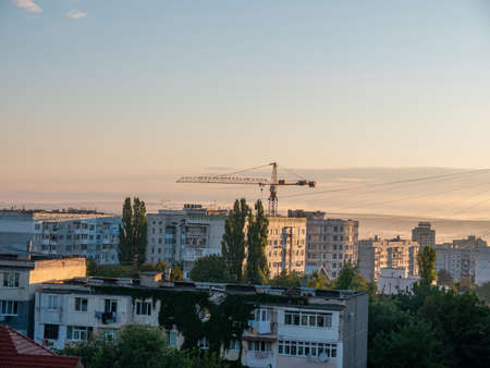 Construction crane over the houses against a blue sky with green leaves of trees in the foreground....