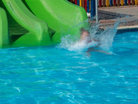 A little girl goes down a green water slide in a pool with blue clear water in a water park. Summer vacation at the resort.の写真素材