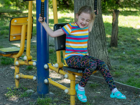 Outdoor sports ground with exercise equipment. A little girl 5-6 years old trains her body muscles...