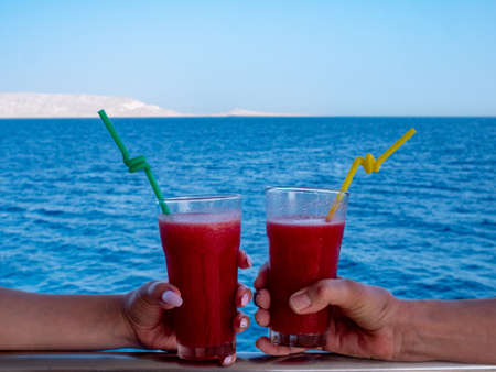 Two female hands hold glasses with fruit cocktails on the background of the sea.の写真素材