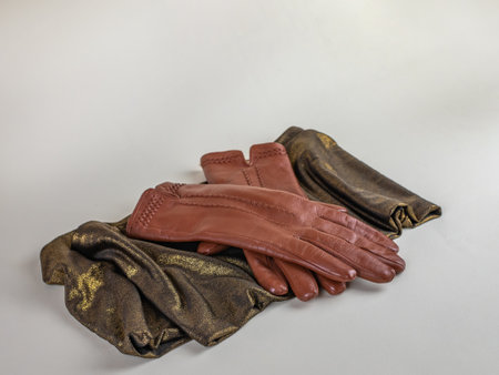 A pair of new brown leather gloves on a green and gold scarf, isolated on a light background. Graceful womens brown leather gloves lying on a green scarf with a golden sheen on a light backgroundの写真素材