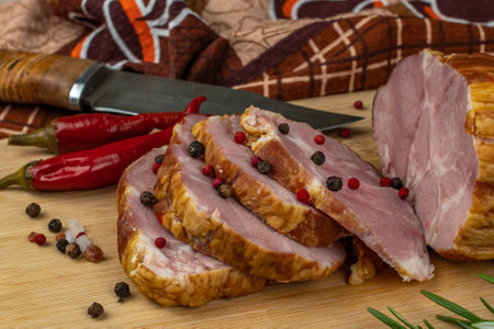 Sliced slices of juicy pork ham on a wooden cutting board, next to it are a carving knife, multi-colored allspice peas, red hot peppers, parsley, dill and basil. The concept of delicious meat productsの写真素材