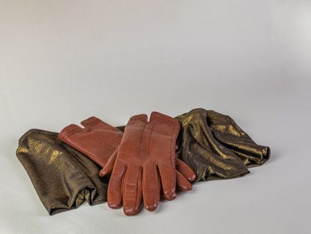 Graceful female brown leather gloves lying on a green scarf with a golden tint on a light background. A pair of new brown leather gloves on a green and gold scarf, isolated on a light backgroundの写真素材