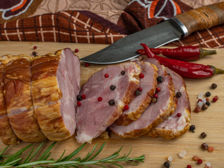 Sliced slices of juicy pork ham on a wooden cutting board, next to it are a carving knife, multi-colored allspice peas, red hot peppers, parsley, dill and basil. The concept of delicious meat productsの写真素材