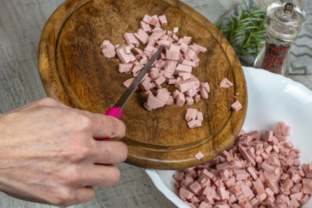Boiled sausage cut into small pieces on a wooden cutting board is poured into a bowl with a knife. Close-up female hands preparing sausage for meat salad. Housewife prepares meat saladの写真素材