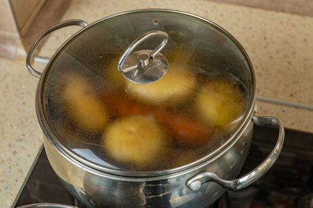 Gas home stove with potatoes and carrots boiling on it in a stainless steel pan without a lid. The concept of cooking healthy food. Close-up.の写真素材