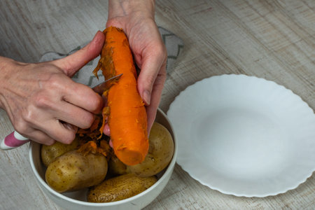 Peeling boiled red carrots. The female hand cuts off the peel from boiled yellow carrots with an iron table knife, puts the peeled carrots in a plate with boiled potatoes. Close-up from the top sideの写真素材
