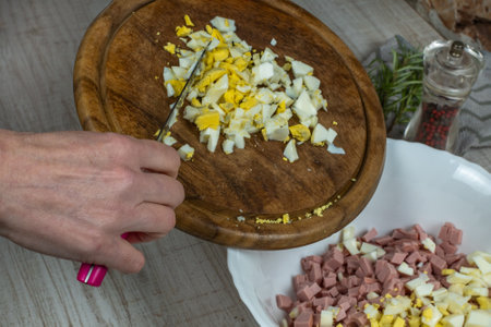 A housewife cuts a boiled chicken egg finely on a wooden cutting board and pushes the egg with a knife into a white glass bowl for making meat salad. Close-up of female hands preparing meat saladの写真素材