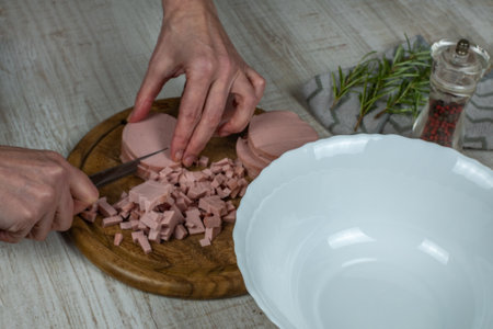 Sausage is cut on wooden cutting board with kitchen knife. Female hands slicing boiled sausage with a knife on a wooden cutting board on a gray kitchen table. Pepper grinder, rosemary sprig. Close-upの写真素材